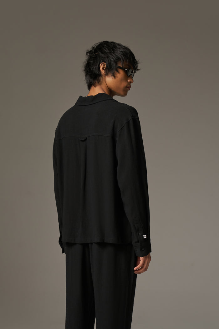 The back image of a man wearing a long sleeved shirt, with black sunglasses and pants against a neutral background.