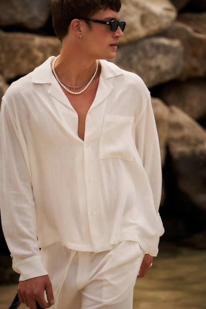 Person wearing a white long sleeved shirt and pants with sunglasses, standing against a stone wall.