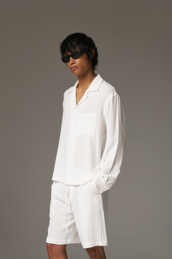 Man wearing a long sleeved white shirt, shorts and sunglasses against a neutral background.