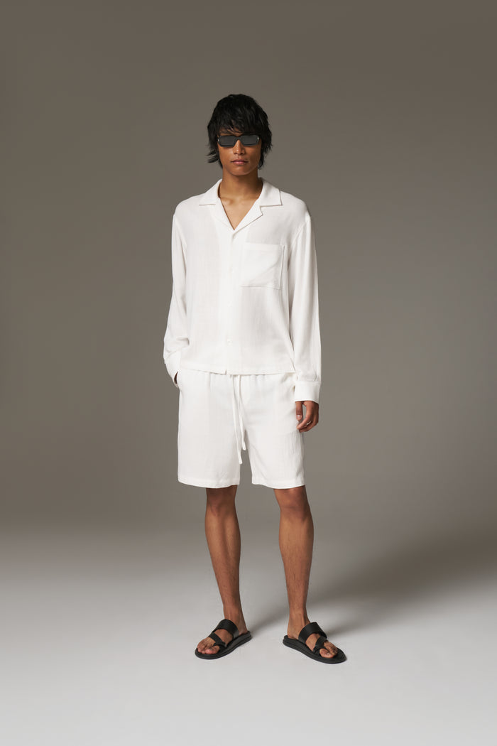 Man wearing a long sleeved white shirt, shorts and sunglasses against a neutral background.