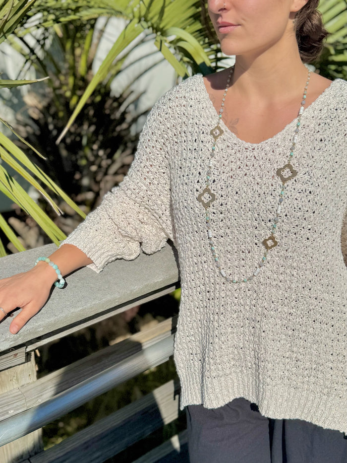 Woman wearing a light-colored crochet top with a necklace, standing outdoors with greenery in the background.