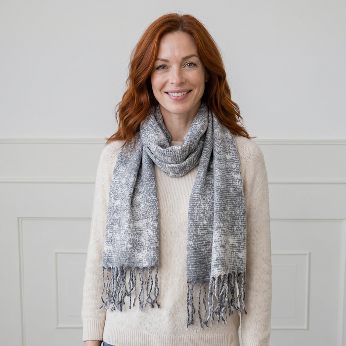 Woman wearing a beige sweater and blue jeans with a gray patterned scarf against a white paneled wall.