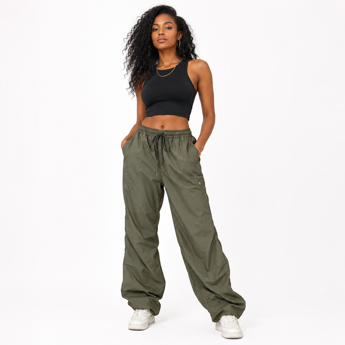 A woman modeling a pair of loose-fitting olive green drawstring pants with a black crop top and white sneakers, standing against a plain white studio background.