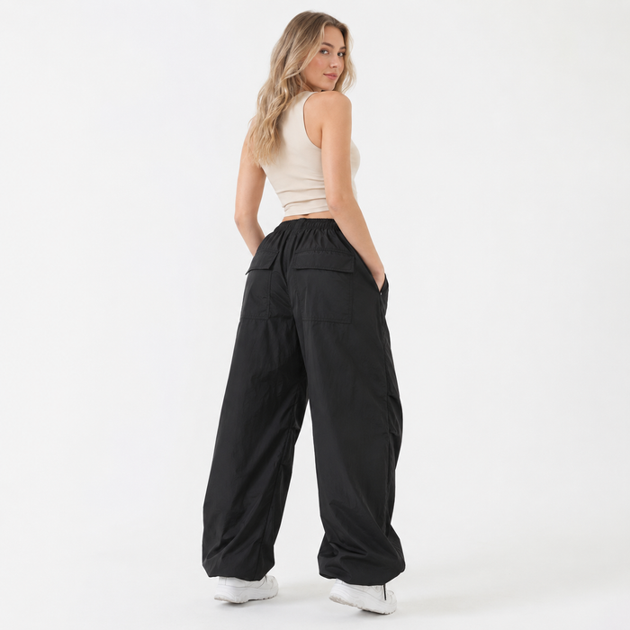 A woman modeling a pair of black wide-leg pants with an elastic waistband and side pockets, paired with a beige crop top and white sneakers, photographed against a solid white studio background.
